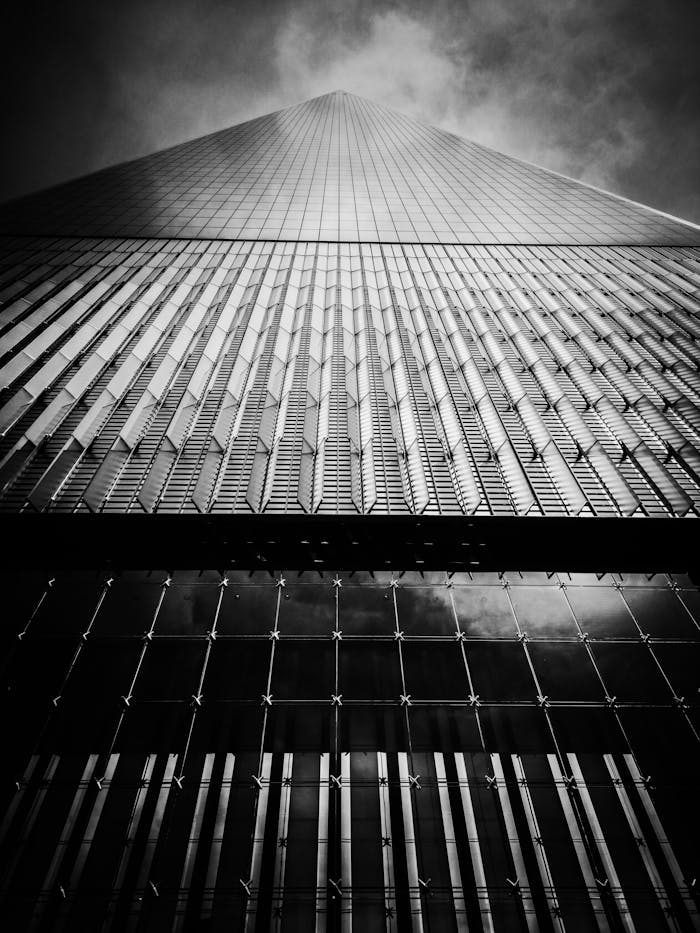 Black and white low angle view of One World Trade Center reaching into the cloudy sky with dramatic architecture.